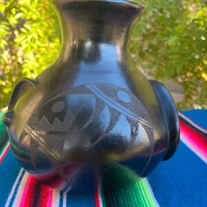 May include: A black ceramic vase with a glossy finish. The vase features a detailed etched design on the front, with a rounded body and a narrow neck. Two small handles are on the sides.