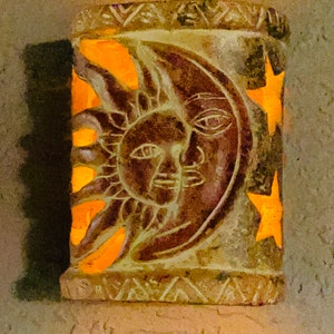 May include: A carved stone candle holder with a sun and moon design. The candle holder is lit and glows a warm orange color.