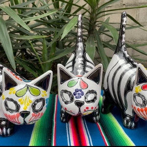 May include: Three ceramic cat figurines with black and white skeletal designs and colorful Day of the Dead inspired patterns. The cats are standing on a colorful striped blanket.