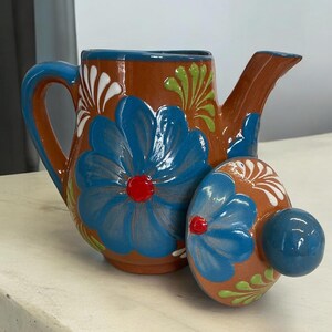 May include: A brown ceramic teapot with a blue and white floral design. The teapot has a lid with a knob on top. The teapot is sitting on a white surface.
