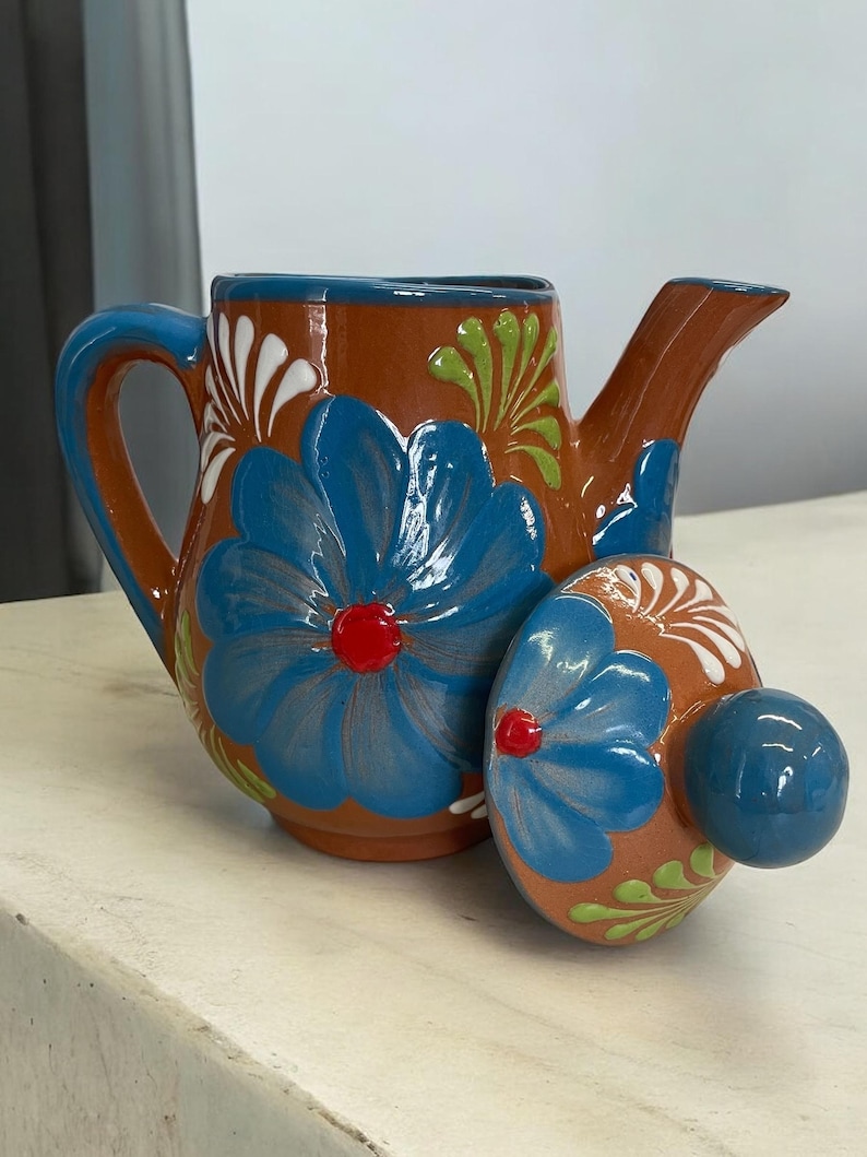 May include: A brown ceramic teapot with a blue and white floral design. The teapot has a lid with a knob on top. The teapot is sitting on a white surface.