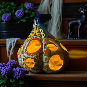 May include: A colorful ceramic pumpkin-shaped lantern with cut-out holes, illuminated from within. The lantern features a dark blue handle and is decorated with floral and geometric patterns in various colors. A black cat figurine and purple flowers are in the background.