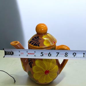 May include: A small orange ceramic teapot with a floral design. The teapot has a lid and a spout. The flowers are yellow with red centers and green leaves. The teapot is 5 inches tall.