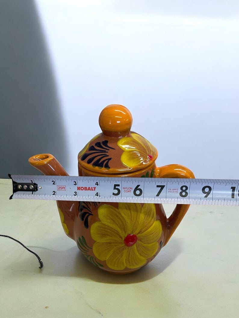 May include: A small orange ceramic teapot with a floral design. The teapot has a lid and a spout. The flowers are yellow with red centers and green leaves. The teapot is 5 inches tall.