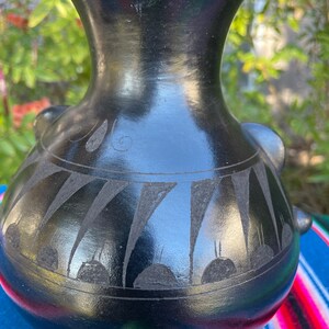 May include: A black ceramic vase with a glossy finish. The vase features etched geometric patterns around the body and small handles. The background is blurred, with green foliage and a colorful striped textile.