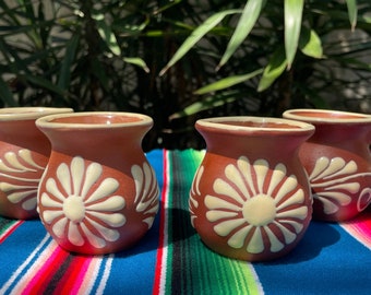 May include: Four terracotta mugs with white floral designs. The mugs have a rounded body and a single handle. They are arranged on a colorful striped blanket.