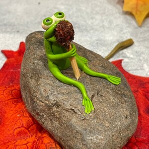 May include: A green frog figurine with large eyes sits on a gray rock, holding a brown and white lollipop in its hands.