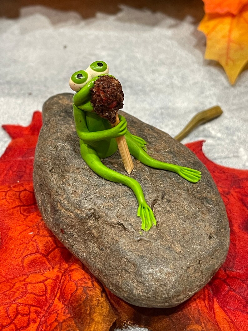 May include: A green frog figurine with large eyes sits on a gray rock, holding a brown and white lollipop in its hands.