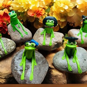 May include: A collection of whimsical green frog figurines, each perched on a stone. The frogs are adorned with various accessories, including hats and headbands. The background features colorful flowers.