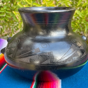 May include: Black ceramic pot with geometric designs carved into the surface. The pot has a rounded body and a wide mouth.