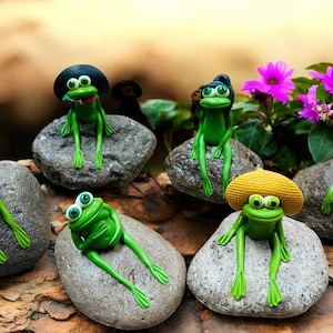 May include: A collection of whimsical green frog figurines, each perched on a gray stone. Some frogs wear hats, and all have large, expressive eyes. The scene is set outdoors with vibrant pink flowers in the background.