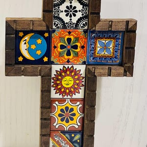 May include: Decorative wooden cross featuring colorful ceramic tiles. The tiles display various designs, including a sun, moon, and floral patterns. The cross is framed with dark brown wood and has a natural fiber loop for hanging.