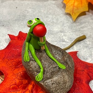May include: A green frog figurine with large eyes sits on a brown rock, holding a red lollipop. The frog is sitting on a red and orange leaf.
