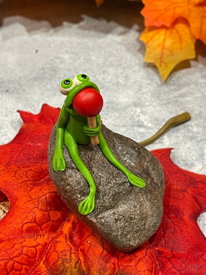 May include: A green frog figurine with large eyes sits on a brown rock, holding a red lollipop. The frog is sitting on a red and orange leaf.