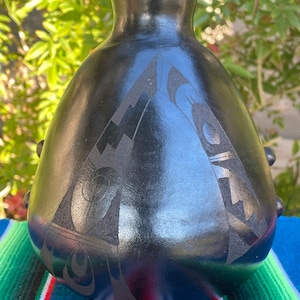 May include: Black ceramic vase with a geometric design. The vase has a rounded body and a narrow neck. The design is carved into the surface of the vase and is a dark gray color.