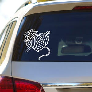 May include: A white vinyl decal applied to the rear window of a vehicle. The design features a ball of yarn shaped like a heart with a crochet hook inserted through it, accompanied by the text "I'd Rather Be Crocheting" in a curved font.