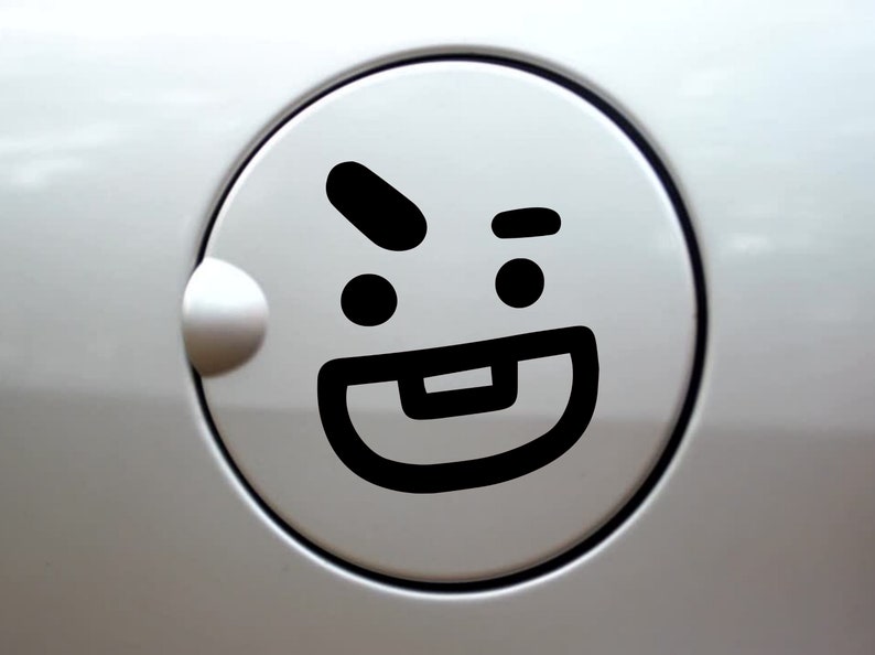 May include: A circular white car fuel door featuring a black vinyl decal of a cartoon face. The graphic depicts a winking eye, a raised eyebrow, and a wide mouth with a single square tooth, creating a playful expression on the smooth surface.
