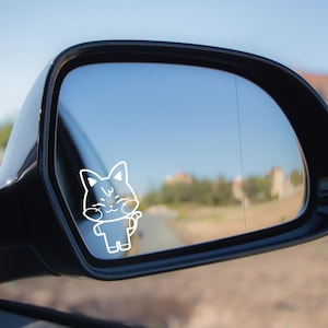 May include: White outline sticker of a cartoon cat waving, applied to a car side mirror. The cat has a smiling face and is in a standing position. The background shows a road and buildings under a blue sky.