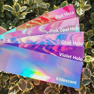 May include: A fan of five rectangular adhesive strips with holographic finishes in red, pink opal, opal, violet, and iridescent colors. Each strip features white text identifying the color: Red Holo, Pink Opal Holo, Opal Holo, Violet Holo, and Iridescent.