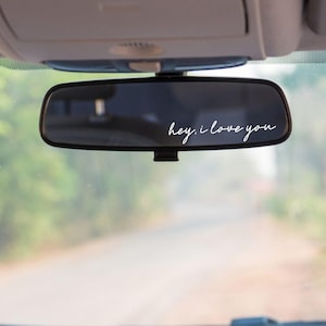 May include: A car's rearview mirror with the phrase "hey, I love you" in white script. The mirror is rectangular and mounted on the windshield. The background is a blurred view of a road and trees.
