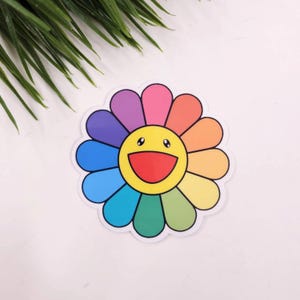 May include: A colorful rainbow flower sticker with a smiling yellow face in the center. The flower has 12 petals and is outlined in black.