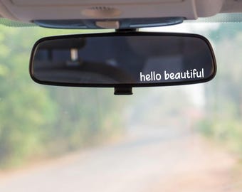 May include: A rectangular car rearview mirror featuring the white text hello beautiful applied to the glass surface. The mirror is mounted to the interior ceiling of a vehicle, reflecting a blurred outdoor scene through the windshield.