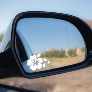 May include: A white hibiscus flower decal is affixed to a black car side mirror. The mirror reflects a clear blue sky and a distant landscape. The decal includes the flower and several falling petals.