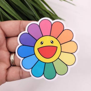 May include: A colorful sticker of a smiling flower with a rainbow of colors. The flower has a yellow center with a smiling face.