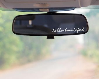 May include: A black car rearview mirror featuring a white vinyl decal with the text hello beautiful written in a cursive script font. The decal is applied to the lower right corner of the mirror surface, which reflects a blurred outdoor road scene.