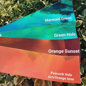 May include: Four rectangular adhesive strips with a reflective finish, arranged in a fan shape. The colors are labeled with white text: Mermaid Green, Green Holo, Orange Sunset, and Peacock Holo Grn/Orange tone. Each strip has a smooth, glossy surface.