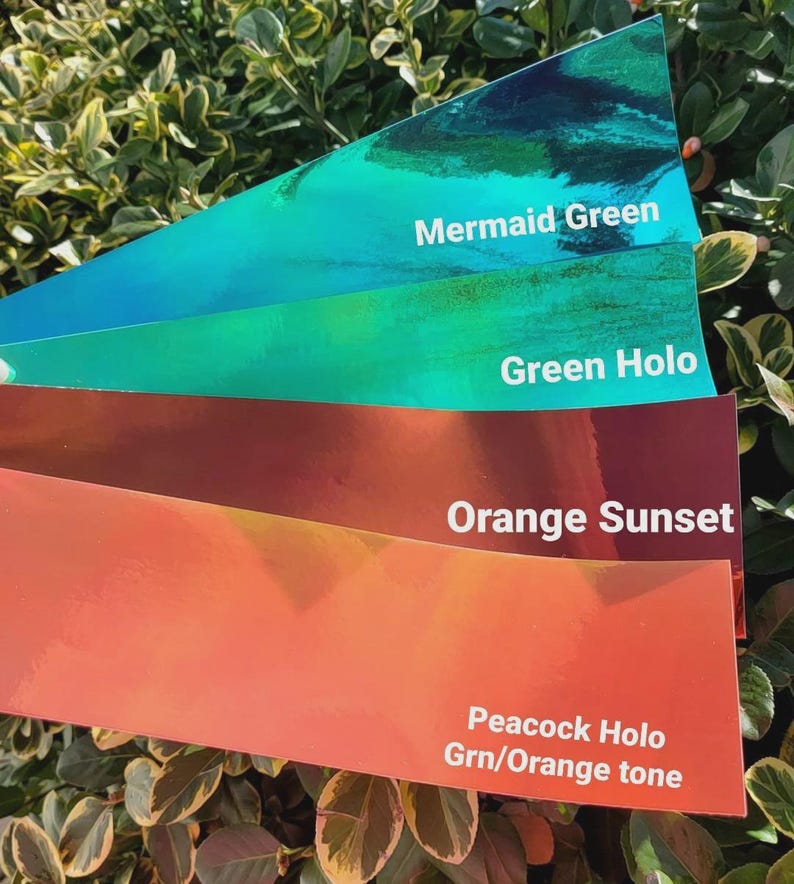 May include: Four rectangular adhesive strips with a reflective finish, arranged in a fan shape. The colors are labeled with white text: Mermaid Green, Green Holo, Orange Sunset, and Peacock Holo Grn/Orange tone. Each strip has a smooth, glossy surface.