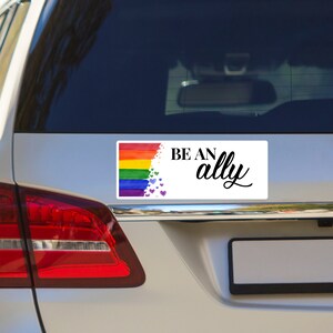 May include: A white car bumper sticker with a rainbow flag and the text "BE AN ally" in black lettering. The rainbow flag is made of watercolor paint and has purple hearts scattered around it.