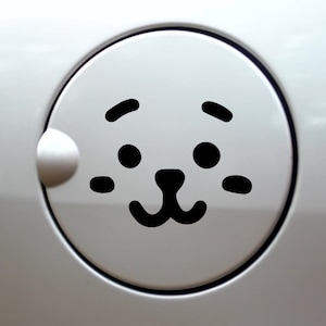 May include: A circular white car fuel door cover featuring a black graphic decal of a stylized animal face. The design includes two round eyes, two small eyebrows, two horizontal cheek marks, and a small nose with a curved mouth centered on the lid.