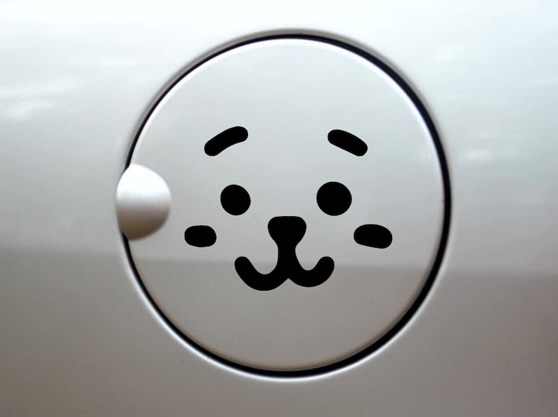 May include: A circular white car fuel door cover featuring a black graphic decal of a stylized animal face. The design includes two round eyes, two small eyebrows, two horizontal cheek marks, and a small nose with a curved mouth centered on the lid.