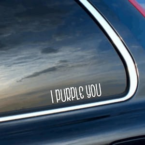 May include: A white vinyl decal sticker applied to a dark car window, featuring the text I PURPLE YOU in a rounded, sans-serif font. The lettering is clean and bold, contrasting against the dark glass surface of the vehicle window.