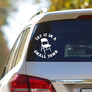 May include: White vinyl sticker with the text "Try it in a small town" in a circle with a folding chair in the center.