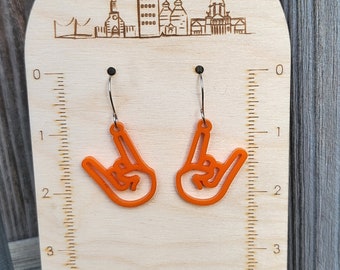 May include: Orange acrylic earrings shaped like a hand giving the rock and roll hand gesture. The earrings are displayed on a wooden stand with a ruler and a city skyline design. The stand is labeled "glo Co & O".