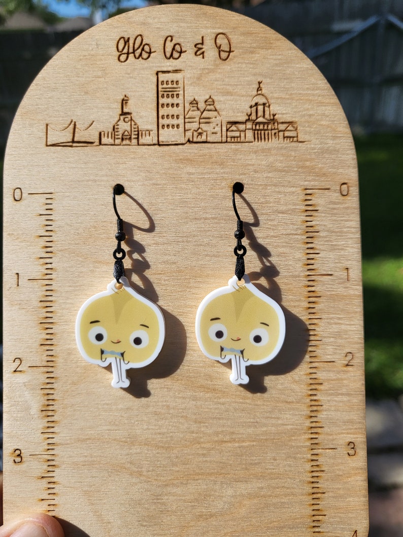Op de afbeelding: Een paar oorhangers met cartoon-ui-figuren met glimlachende gezichten. De oorhangers zijn gemaakt van wit acryl en hebben zwarte haakjes.