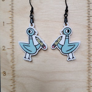 Op de afbeelding: Een paar oorbelhangers met een cartoon blauwe vogel die een potlood in zijn snavel vasthoudt. De oorringen hebben zwarte haken en zijn gemaakt van acryl.