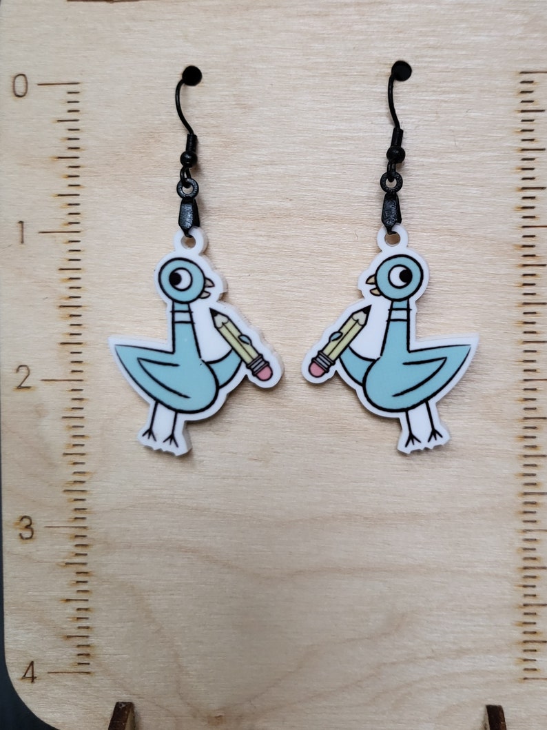 Op de afbeelding: Een paar oorbelhangers met een cartoon blauwe vogel die een potlood in zijn snavel vasthoudt. De oorringen hebben zwarte haken en zijn gemaakt van acryl.