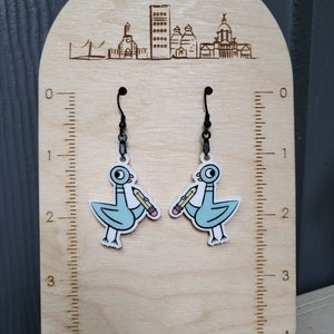 Op de afbeelding: Een paar hangende oorbellen met een lichtblauwe cartoonduif die een geel potlood vasthoudt. De oorbellen worden getoond op een houten standaard met een liniaal en de tekst "Glo Co & D" gegraveerd aan de bovenkant.