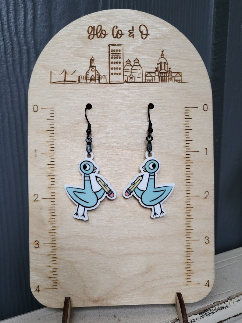 Op de afbeelding: Een paar hangende oorbellen met een lichtblauwe cartoonduif die een geel potlood vasthoudt. De oorbellen worden getoond op een houten standaard met een liniaal en de tekst "Glo Co & D" gegraveerd aan de bovenkant.