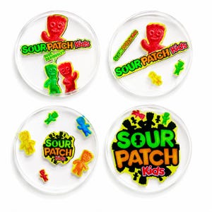 以下が含まれることがあります： 「Sour Patch Kids」のロゴとキャンディーのキャラクターが描かれた、透明な丸いコースター4枚。各コースターは異なるデザインで、ロゴは緑とオレンジ、キャンディーは赤、緑、黄色、オレンジです。
