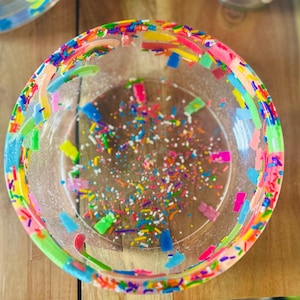 May include: A clear, round bowl with a colorful, confetti-like design. The rim is decorated with a vibrant mix of pink, yellow, green, and blue rectangular shapes. The interior is filled with tiny, multicolored sprinkles.