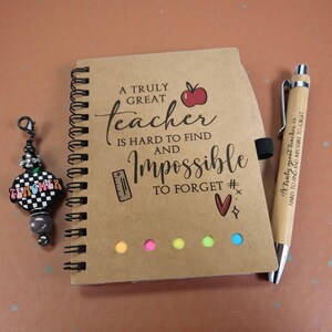 May include: A spiral-bound notebook with the text "A Truly Great Teacher Is Hard to Find and Impossible to Forget." A wooden pen with the same quote is next to the notebook. A charm with the word "Teacher" is also in the image.