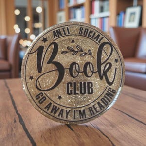 May include: A round, glittery gold Book Club sign with black text. The sign reads "ANTI-SOCIAL Book CLUB GO AWAY I'M READING" in a decorative font. The sign has a gold glitter background and is on a wooden surface.