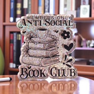 May include: Glittery, layered book club sign with the words "Members Only Anti Social Book Club." The sign features a stack of books, a coffee cup, flowers, and hearts. The sign is on a table in front of a bookshelf.