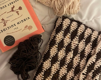 May include: A handmade crochet book cover in brown and cream diamond pattern, with a button closure. Beside it are balls of cream and brown yarn and a copy of "The Virgin Suicides" by Jeffrey Eugenides.