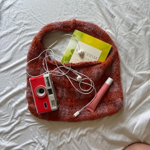 Op de afbeelding: Een gebreide, oranje en rode tas met een boek, een rode camera, oortelefoontjes en een roze lipgloss. De tas ligt op een wit oppervlak.