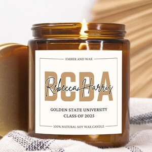 May include: A lit amber glass candle with a gold lid, resting on a striped cloth. The label reads "BCBA Rebecca Harris Golden State University Class of 2025 100% Natural Soy Wax Candle."